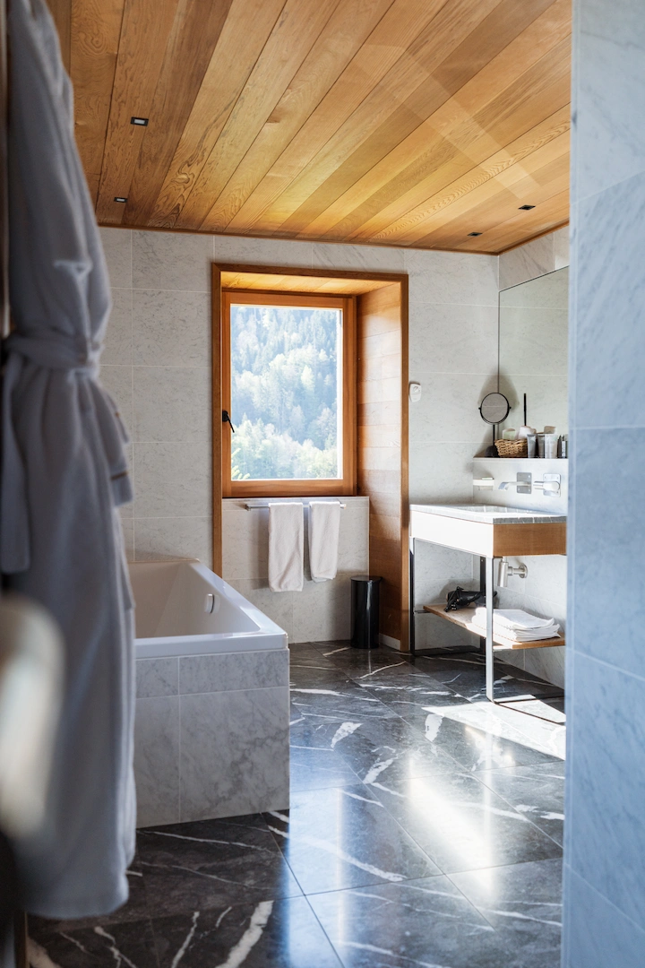Salle de bain élégante en chalet alpin avec baignoire encastrée, sol en marbre noir veiné et plafond en bois clair. Fenêtre à encadrement bois avec vue sur la forêt de montagne, vasque suspendue avec miroir, serviettes et peignoir blanc. Lumière naturelle abondante.