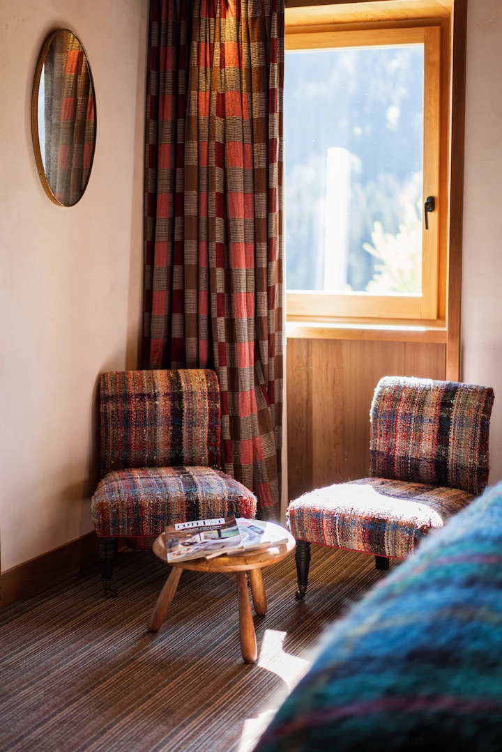 Coin salon d'une chambre d'hôtel chalet avec deux fauteuils en tissu tweed multicolore encadrant une petite table basse en bois avec des magazines. Rideau à carreaux rouge et brun, fenêtre en bois laissant entrer la lumière naturelle, miroir ovale doré au mur. Ambiance chaleureuse et colorée.