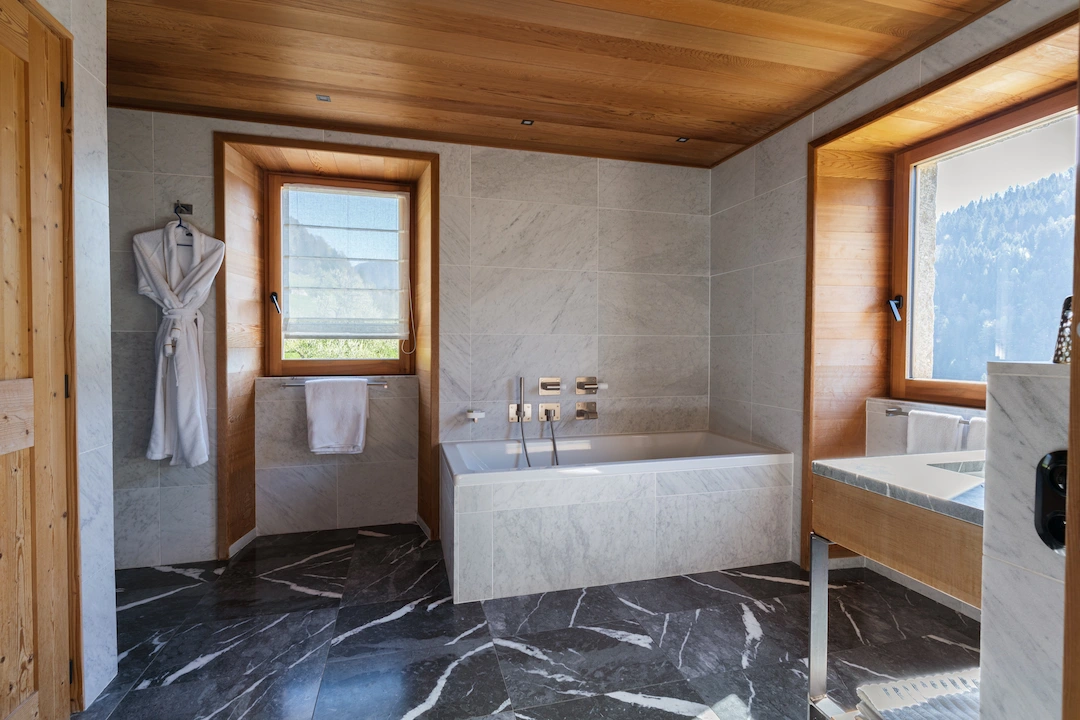 Salle de bain haut de gamme en chalet alpin, avec baignoire encastrée habillée de marbre clair, sol en marbre noir veiné et plafond en bois. Fenêtres à encadrements en bois offrant une vue sur les montagnes. Peignoir blanc suspendu et vasque en marbre visible sur la droite.
