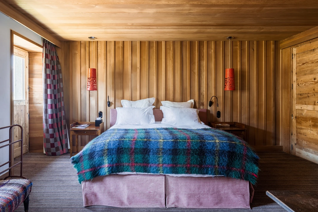 Chambre d'hôtel chalet au style coloré et chaleureux, avec un lit double recouvert d'un plaid en laine à carreaux bleu et vert. Murs et plafond en lambris de bois clair, deux suspensions design orange de chaque côté du lit, rideaux à motif pied-de-poule rouge et gris. Ambiance montagne contemporaine et vivante.