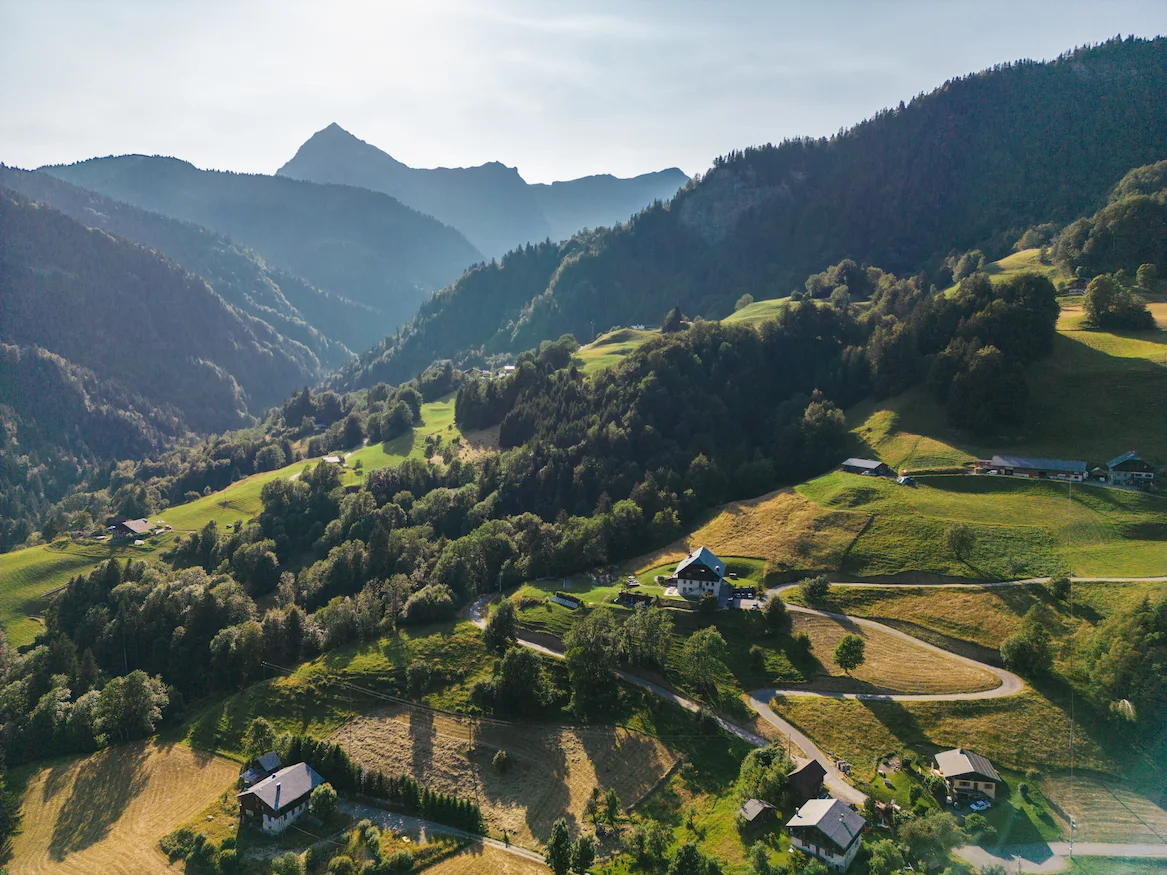Vue aérienne d'une vallée alpine en été avec prairies verdoyantes, forêts de sapins et chalets dispersés sur les flancs. Route sinueuse traversant le paysage, sommet rocheux pointu en arrière-plan dans une lumière dorée de fin de journée. Environnement naturel préservé des Alpes françaises.