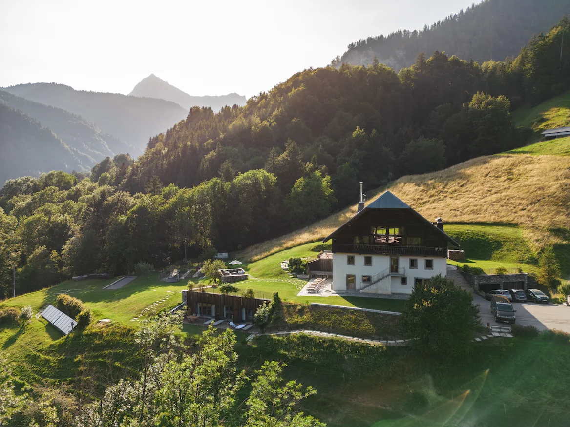 Vue drone d'un domaine alpin en été avec un grand chalet à façade blanche et toit en ardoise, entouré de prairies verdoyantes en pente. Espace spa semi-enterré avec panneaux solaires, terrasse, jardins aménagés et parking visible. Forêts de sapins et chaîne de montagnes baignées d'une lumière dorée en arrière-plan.