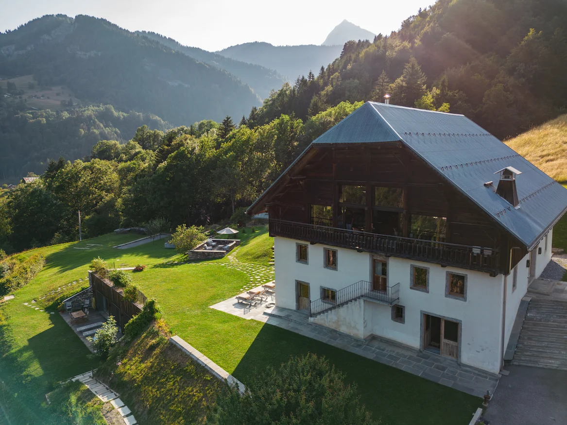 Vue aérienne d'un grand chalet alpin réhabilité à façade blanche et toit en ardoise, avec balcon en bois, terrasse et vaste jardin en pente. Espace spa semi-enterré avec toit végétalisé visible sur la gauche, salon de jardin et parasol. Forêts et sommets montagneux en arrière-plan dans une lumière dorée de fin de journée.