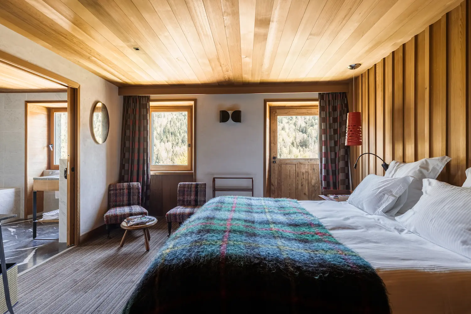 Chambre d'hôtel de montagne avec lit double aux draps blancs recouvert d'un plaid à carreaux bleu et multicolore, tête de lit en lambris bois vertical, deux fauteuils chinés face à deux fenêtres avec vue sur une forêt, lampe suspendue rouge, miroir ovale et accès visible à la salle de bain