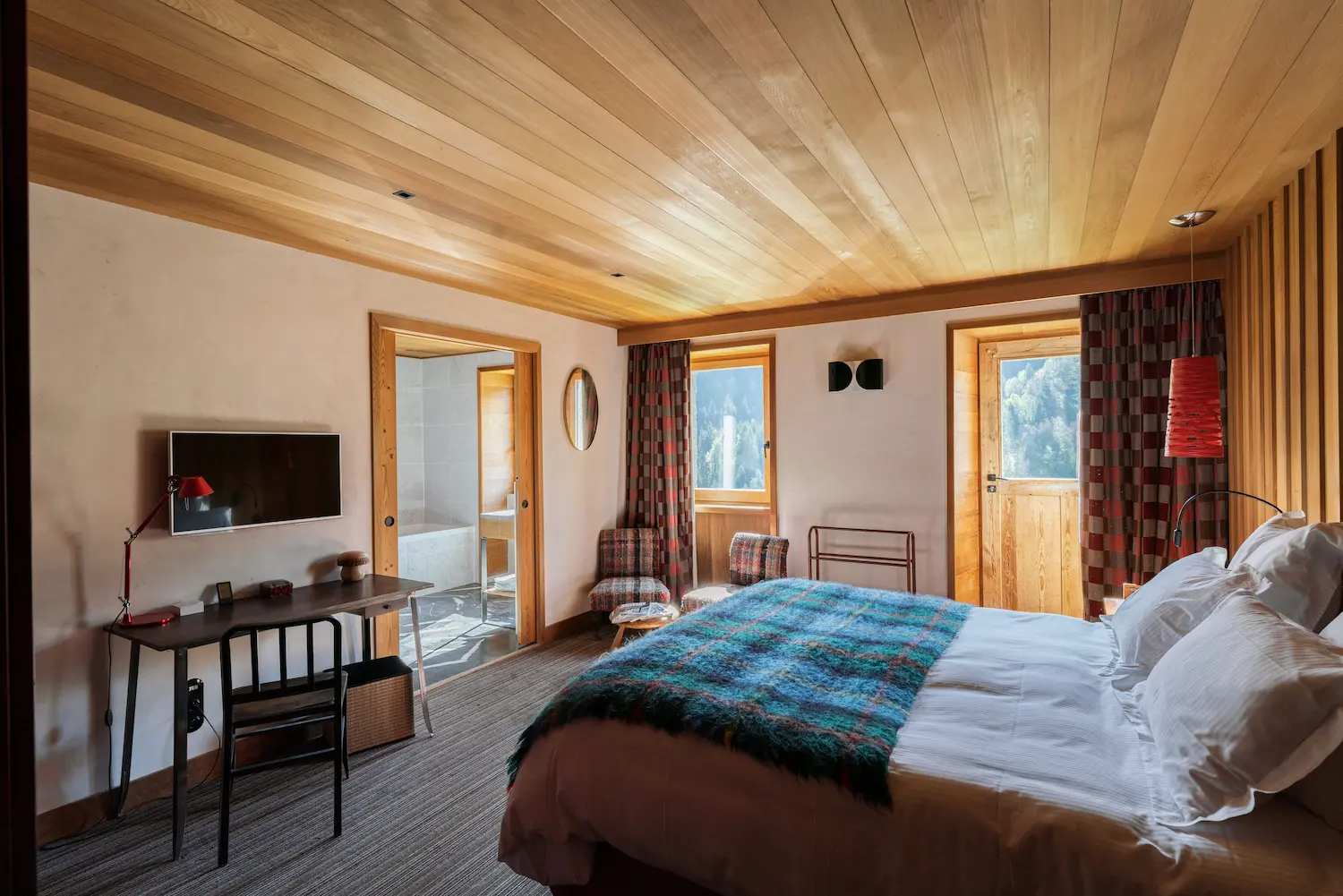 Chambre d'hôtel chaleureuse avec plafond en bois clair, lit double aux draps blancs et plaid bleu, bureau en bois sombre avec télévision murale, fauteuil à carreaux, rideaux à motifs rouges et vue sur une forêt par deux fenêtres