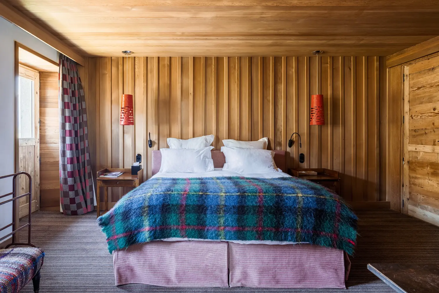 Chambre d'hôtel chalet au style vivant et coloré avec lit double recouvert d'un plaid en laine mohair à carreaux bleu, vert et rouge. Murs en lambris vertical clair, deux suspensions design orange, cache-sommier rose, rideaux à damier rouge et gris. Tables de nuit en bois et petit fauteuil tweed multicolore au premier plan.