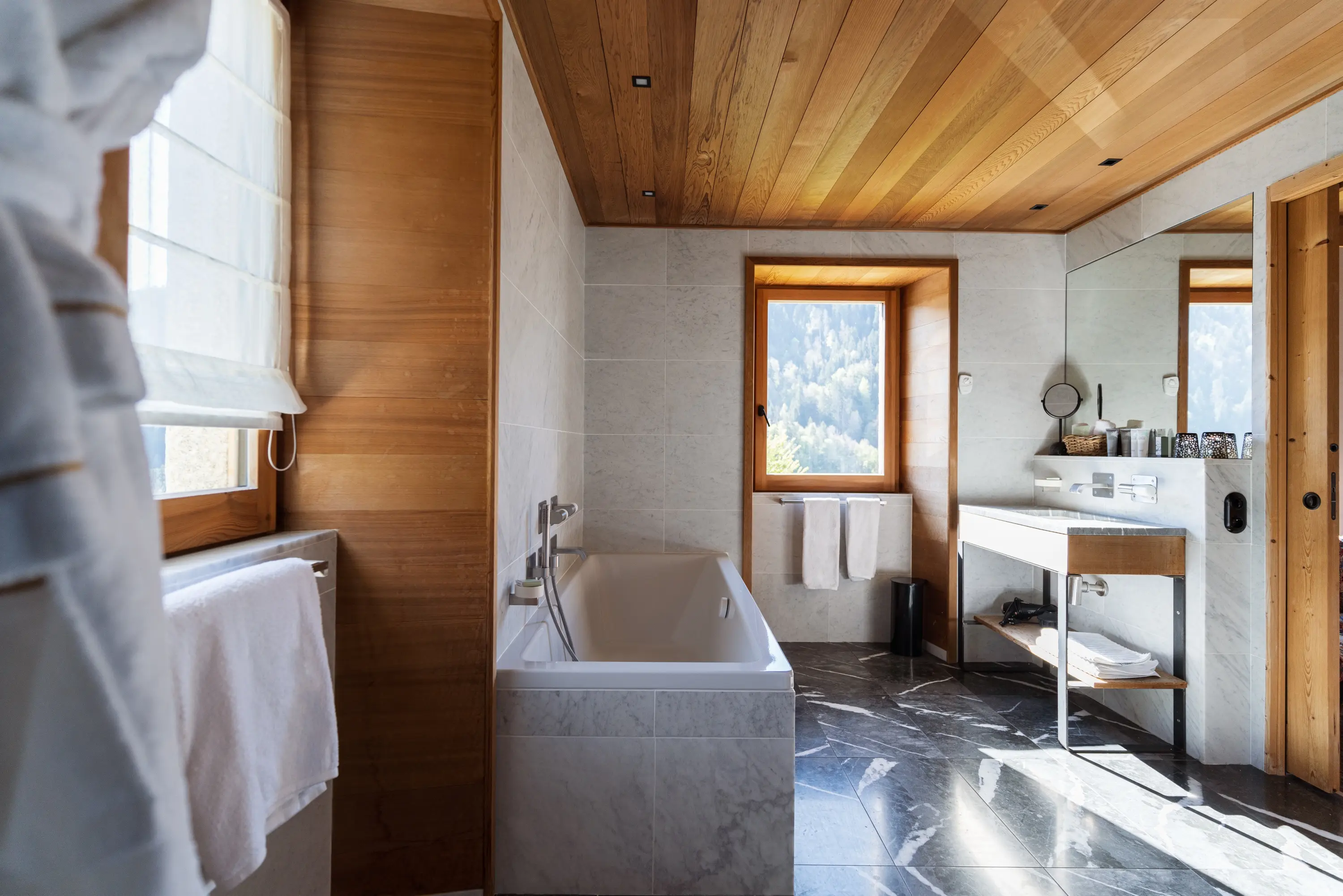Grande salle de bain lumineuse en chalet alpin avec baignoire encastrée en marbre blanc, sol en ardoise noire veinée et plafond en bois clair. Deux fenêtres à encadrement bois avec vue sur la forêt, vasque sur meuble bois et métal avec produits de soin, grand miroir et serviettes blanches. Ambiance spa haut de gamme.