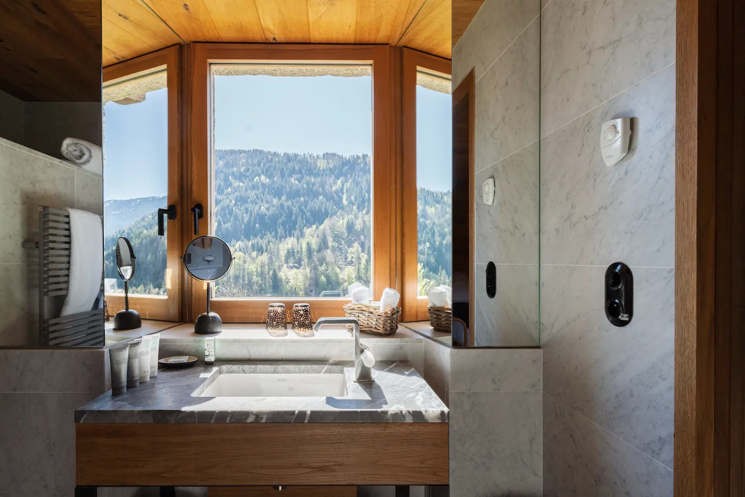 Vasque en marbre gris veiné sur meuble bois avec robinetterie noire, miroir de courtoisie, produits de soin et panier en osier, devant une grande fenêtre à encadrement bois offrant une vue panoramique sur une forêt de montagne et un ciel bleu, avec entrée de douche en marbre blanc visible à droite