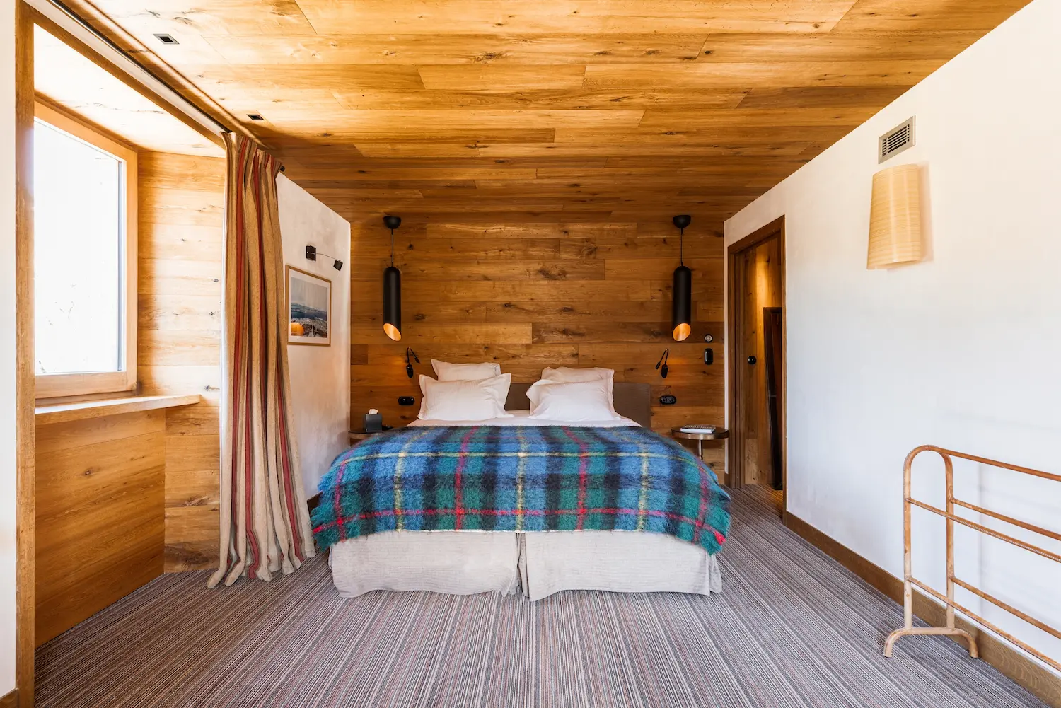 Chambre d'hôtel de montagne avec lit double aux draps blancs recouvert d'un plaid à carreaux bleu et multicolore, tête de lit en lambris bois massif, deux suspensions cylindriques noires, liseuses murales orientables, rideau rayé beige et rouge à gauche, porte-bagages en métal cuivré et plafond en bois clair