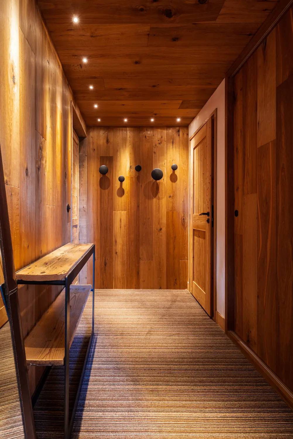 Couloir d'entrée d'hôtel entièrement habillé de bois brut aux teintes chaudes, avec quatre patères rondes noires alignées sur le mur du fond, console fine en métal et bois à gauche, spots encastrés au plafond, moquette rayée au sol et porte en bois sur la droite