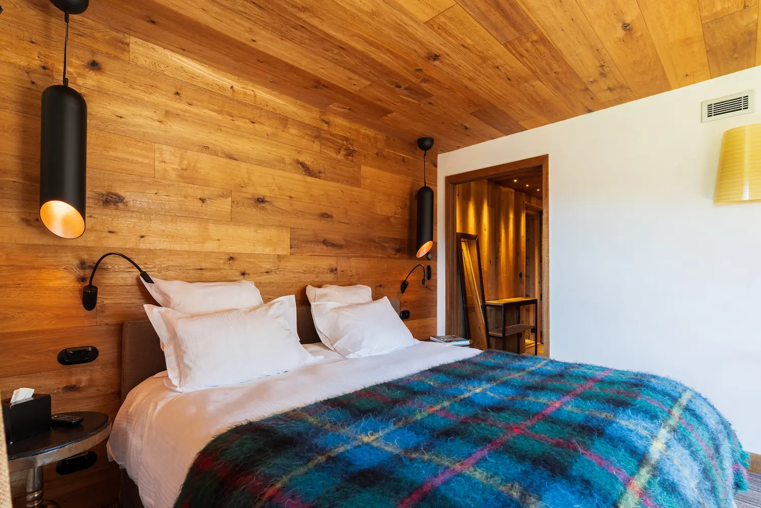 Chambre d'hôtel avec lit double aux draps blancs recouvert d'un plaid à carreaux bleu et multicolore, tête de lit et mur en lambris bois massif, deux suspensions cylindriques noires, liseuses murales orientables et accès visible à un couloir d'entrée en bois chaleureusement éclairé