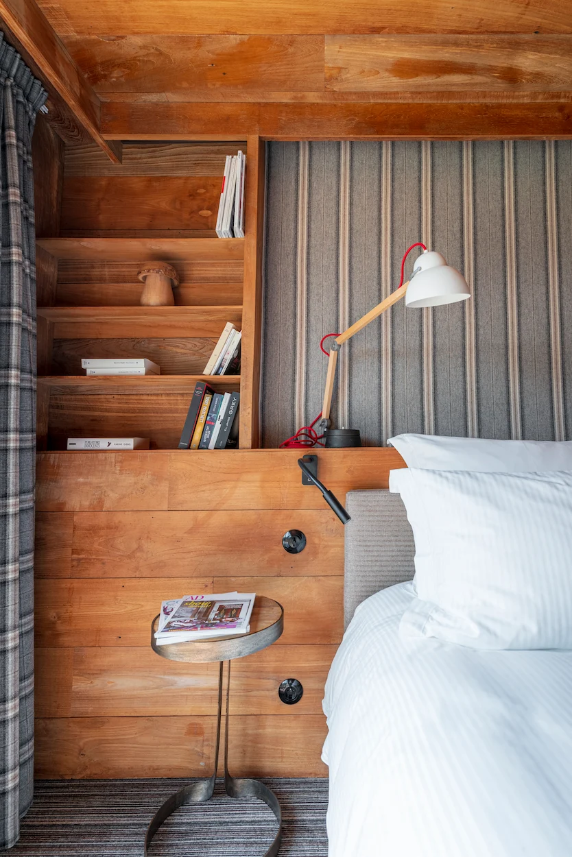 Détail de chevet d'une chambre chalet : bibliothèque intégrée en bois avec livres et magazines, liseuse design blanc et rouge sur fil tressé, petite table d'appoint en métal forgé avec un magazine AD. Tête de lit tapissée de tissu rayé beige, linge blanc et rideau à carreaux gris.