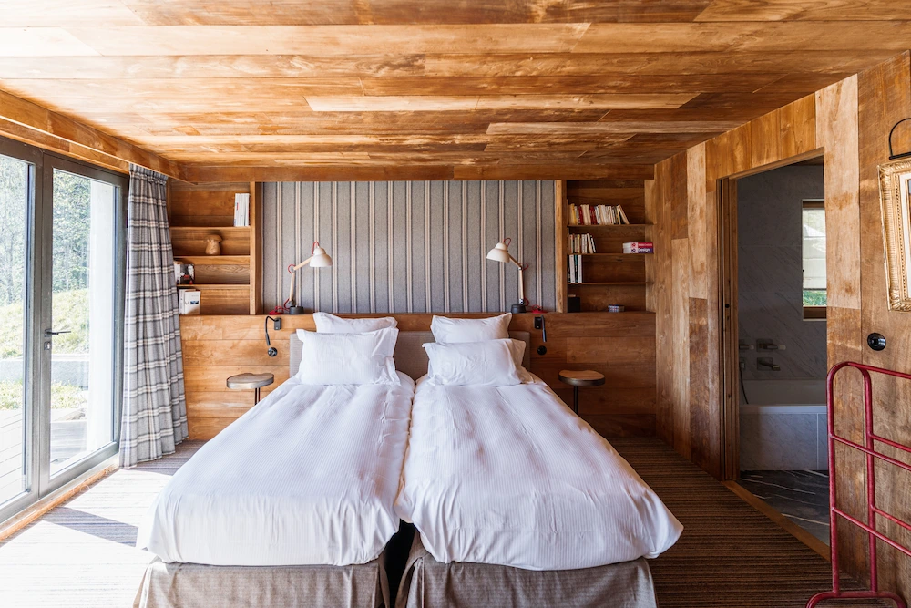 Chambre twin en chalet alpin entièrement habillée de bois clair, avec deux lits en linge blanc, tête de lit tapissée de tissu rayé beige, bibliothèques intégrées et liseuses design. Grande baie vitrée avec rideau à carreaux, salle de bain visible en enfilade et porte-bagages rouge. Ambiance chaleureuse et lumineuse.