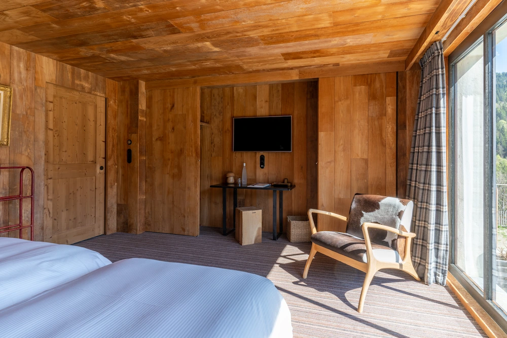Vue d'ensemble d'une chambre d'hôtel entièrement habillée de bois clair, avec lit double, fauteuil vintage en bois recouvert d'une peau de vache, télévision murale et bureau noir. Grande baie vitrée avec rideau à carreaux gris donnant sur la forêt. Porte-bagages rouge visible sur la gauche. Ambiance chalet contemporain baigné de lumière naturelle.