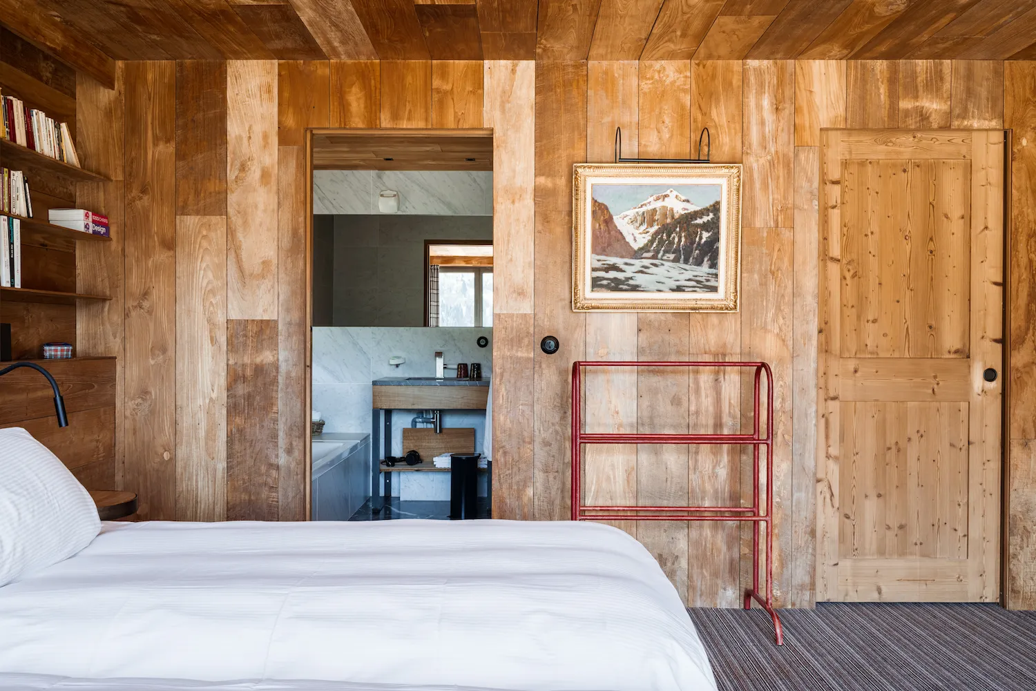 Chambre d'hôtel chalet avec murs en lambris de bois clair, lit double en linge blanc, bibliothèque intégrée et porte-bagages en métal rouge. Tableau ancien représentant un paysage de montagne enneigé accroché au mur. Salle de bain en marbre visible en enfilade. Ambiance montagne contemporaine et raffinée.