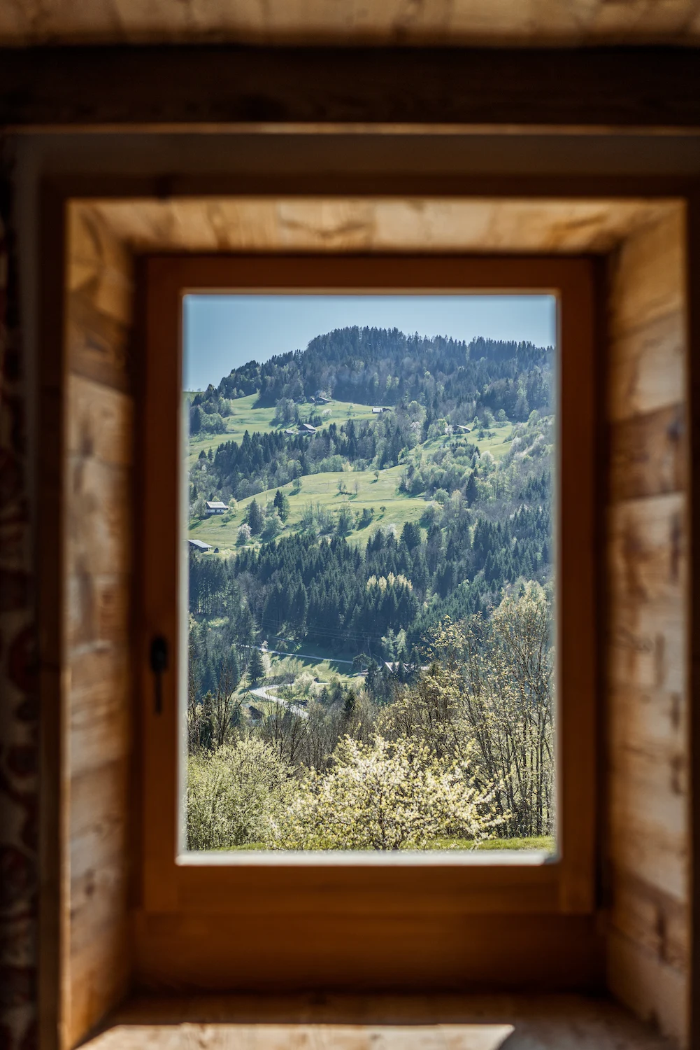 Vue depuis l'intérieur d'un chalet à travers une fenêtre à encadrement en bois sur un paysage alpin printanier : prairies verdoyantes, forêts de sapins, chalets dispersés sur les flancs et ciel bleu. Composition encadrée et lumineuse évoquant l'invitation à la montagne.