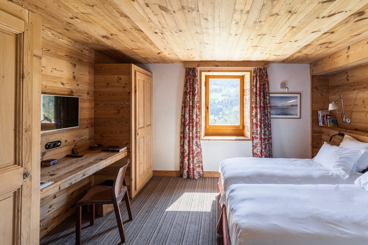 Chambre twin en chalet alpin avec deux lits simples en linge blanc, murs et plafond en bois de pin clair. Bureau intégré avec télévision, fenêtre à encadrement bois avec rideaux à motifs floraux rouges donnant sur une forêt. Photo de paysage et livres sur une étagère côté lit.