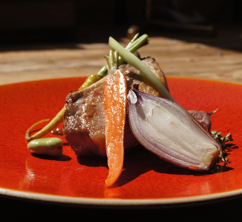 Assiette gastronomique sur céramique rouge avec une pièce de viande rôtie accompagnée de légumes du marché : carotte glacée, oignon rouge confit, haricots verts et herbes fraîches. Dressage soigné et cuisine bistronomique.