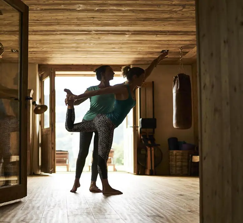 Coach guidant une cliente dans une posture d'équilibre de yoga dans une salle de sport en chalet alpin. Plafond et sol en bois clair, sac de frappe suspendu en arrière-plan, baie vitrée ouvrant sur un paysage de montagne. Lumière naturelle en contre-jour.