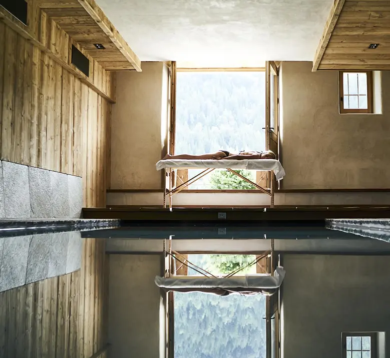 Personne allongée sur une table de massage au bord d'une piscine intérieure, face à une grande fenêtre ouverte sur une forêt de montagne. Le reflet symétrique de la scène dans l'eau calme de la piscine crée une composition épurée. Murs en bois et enduit naturel, ambiance spa alpin serein.