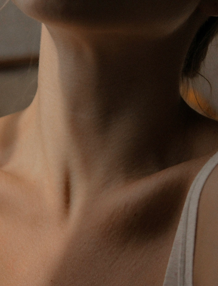 Gros plan sur le cou et le décolleté d'une femme en brassière blanche, éclairés par une lumière douce et contrastée. Évocation sensorielle du bien-être, de la détente et du soin corporel.