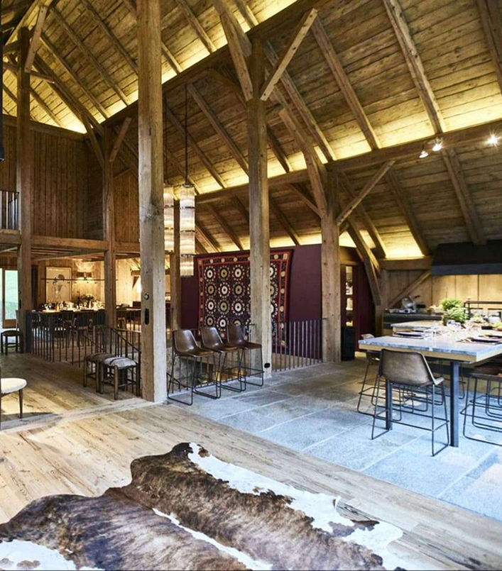 Grand espace de vie ouvert dans un chalet alpin réhabilité, avec charpente apparente en bois massif à haute voûte. Au premier plan, une peau de vache en tapis et une grande table à manger. Au centre, des tabourets de bar face à un panneau décoratif géométrique bordeaux. En fond, un espace restaurant avec tables dressées. Sol mixte bois et pierre.