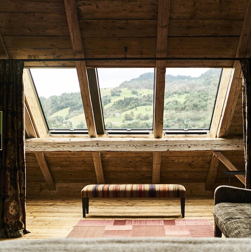 Espace sous combles d'un chalet alpin en bois ancien avec trois grandes fenêtres de toit offrant une vue panoramique sur des prairies verdoyantes et des collines boisées. Banquette à rayures multicolores, tapis rose terracotta et plancher en vieux bois. Ambiance rustique et lumineuse.