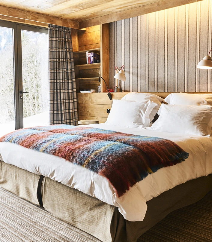 Chambre d'hôtel chalet avec lit double en linge blanc et jeté en laine mohair multicolore aux tonalités rouge, bleu et jaune. Tête de lit tapissée de tissu rayé beige, liseuses design blanches, niches en bois avec livres et baie vitrée donnant sur une forêt enneigée. Ambiance cosy et colorée.
