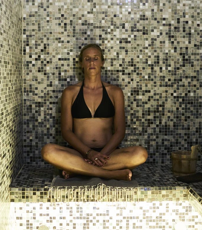 Femme en bikini noir assise en posture de méditation dans un hammam carrelé de mosaïques beige et gris, yeux fermés, mains posées sur les genoux. Seau en bois visible à droite. Ambiance spa et détente profonde.
