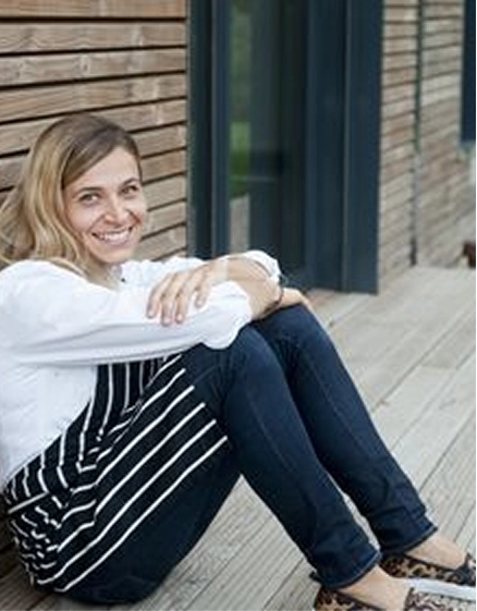 Portrait d'une femme souriante assise sur une terrasse en bois, vêtue d'une chemise blanche et d'un jean, devant une façade en bois et une baie vitrée.