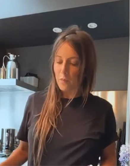 Femme aux cheveux longs en t-shirt gris debout dans une cuisine moderne, regard baissé. Étagères avec ustensiles et carafe en inox visibles en arrière-plan.
