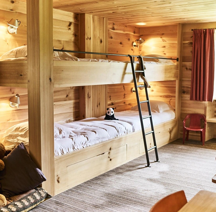 Chambre enfants en chalet alpin avec lits superposés en bois clair intégrés, échelle métallique noire et liseuses orientables. Peluche panda posée sur le lit du bas, peluche ours au sol, chaise rouge et rideau bordeaux. Ambiance chaleureuse et cosy adaptée aux familles.