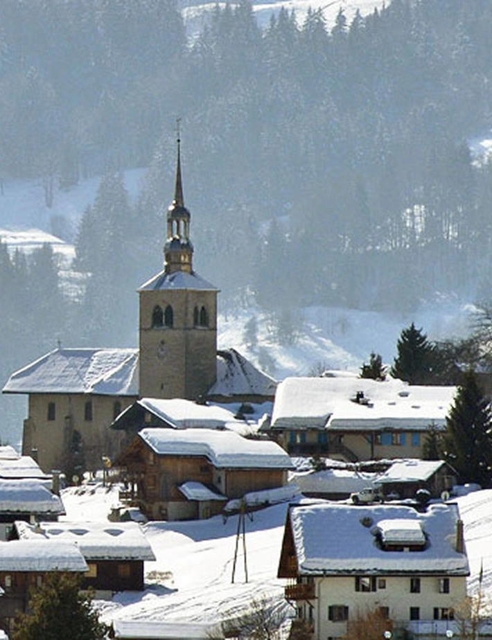 Vue du village de Morzine en hiver sous la neige, avec l'église et son clocher baroque doré au centre, entourée de chalets aux toits enneigés. Forêt de sapins et pistes de ski en arrière-plan dans une lumière hivernale bleutée.