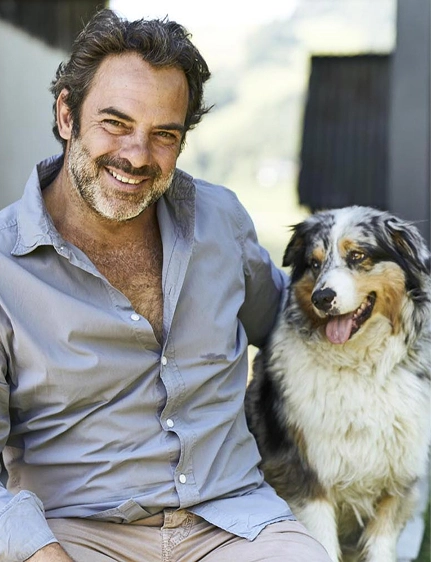 Portrait d'un homme souriant en chemise bleue posant aux côtés d'un berger australien tricolore, en extérieur avec un arrière-plan flou.