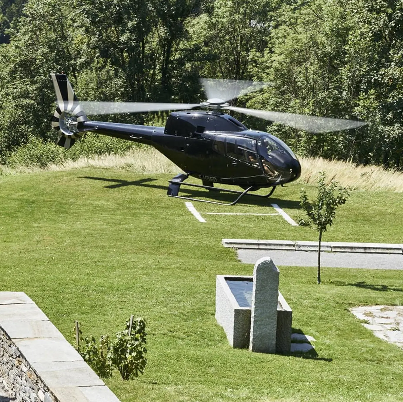 Hélicoptère privé se posant sur l'hélisurface engazonnée du domaine, entouré de verdure.