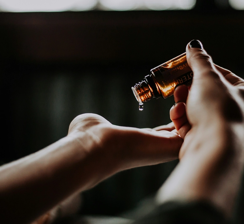Gros plan sur deux mains, l'une versant une goutte d'huile essentielle depuis un flacon en verre ambré sur le poignet de l'autre. Ambiance sombre et intimiste évoquant un rituel de bien-être ou d'aromathérapie.