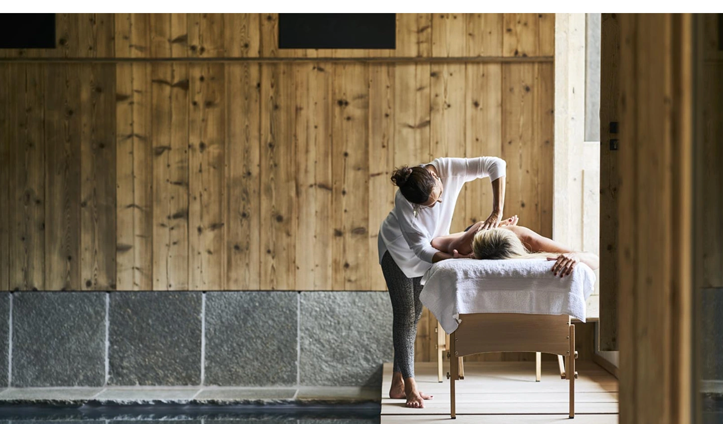 Praticienne réalisant un massage bien-être sur une table de soin, dans un spa en bois attenant à une piscine intérieure.