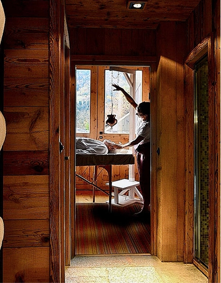 Vue depuis un couloir en bois sur une cabine de soin spa. Un thérapeute prodigue un soin visage à un client allongé sur une table de massage, avec un dispositif de shirobhyanga suspendu au plafond. Lumière naturelle entrant par une baie vitrée avec vue sur la forêt.