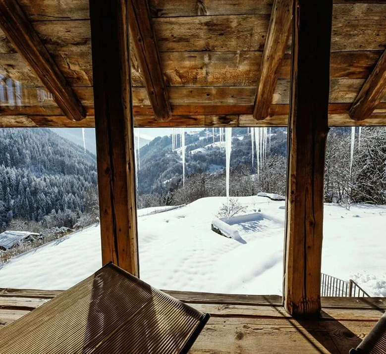 Vue depuis l'intérieur d'un chalet alpin en bois ancien, à travers une grande baie vitrée avec des stalactites de glace, sur un paysage hivernal enneigé avec forêt de sapins et vallée de montagne. Chaise longue en bois au premier plan. Ambiance cosy et contemplative.