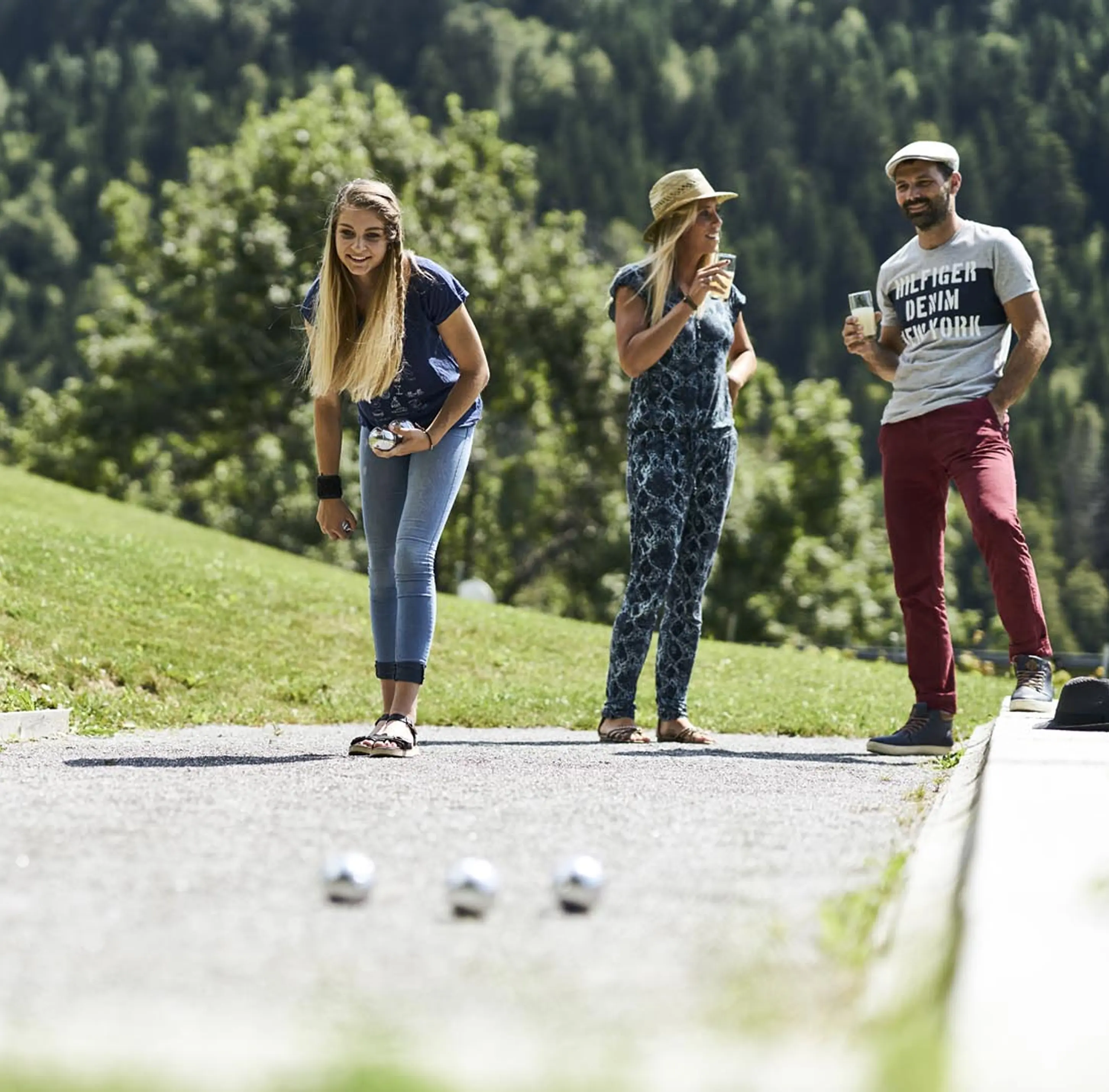 Trois personnes jouant à la pétanque en plein air dans un cadre alpin ensoleillé. Une jeune femme s'apprête à lancer sa boule tandis que deux autres regardent en souriant, verres à la main. Prairie verdoyante et forêt de sapins en arrière-plan.