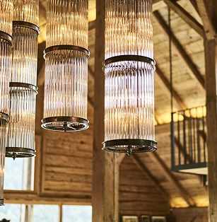 Gros plan sur des suspensions design en verre cannelé et métal doré suspendues au plafond d'un chalet alpin à charpente apparente. Lumière chaude et ambrée, escalier en colimaçon visible en arrière-plan.