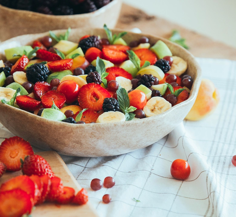 Grande salade de fruits frais dans un bol en céramique beige : fraises, mûres, banane, raisin, melon et feuilles de menthe. Fruits éparpillés autour sur un torchon blanc. Présentation colorée et généreuse évoquant une alimentation saine et gourmande.