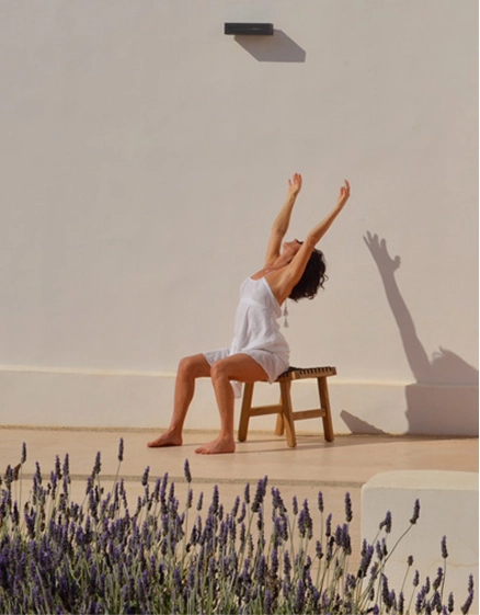 Femme en robe blanche assise sur un tabouret en bois, bras levés vers le ciel dans un mouvement d'étirement ou de yoga, en extérieur face à un mur blanc baigné de soleil. Plants de lavande en fleurs au premier plan, ombre portée des bras sur le mur.