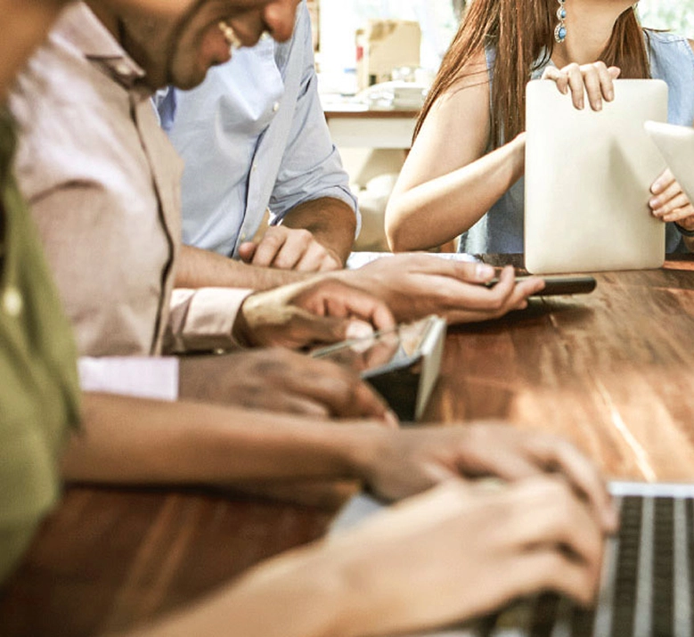 Groupe de professionnels travaillant côte à côte autour d'une table en bois, avec laptops, tablette et smartphone. Ambiance réunion de travail collaborative et détendue en espace de coworking ou salle de séminaire.