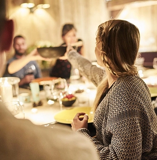 Groupe de personnes réunies autour d'une grande table pour un dîner convivial en intérieur, éclairage tamisé et chaleureux. Au premier plan, une femme aux tresses tend un plat à travers la table. Ambiance repas partagé et convivialité alpine.