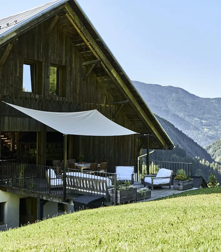 Terrasse en bois d'un chalet alpin avec voile d'ombrage blanche, salon de jardin et table à manger extérieure, surplombant une vallée de montagne. Prairie verte au premier plan, panorama de sommets alpins boisés sous un ciel bleu dégagé.