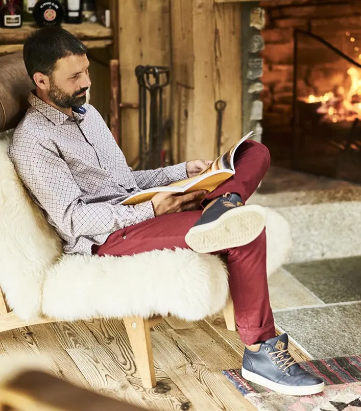Homme lisant un magazine installé dans un fauteuil recouvert d'une peau de mouton blanche, devant une cheminée en pierres allumée. Intérieur chalet alpin en bois, ambiance chaleureuse et détendue après-ski.