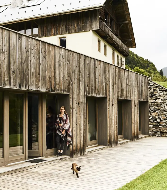 Femme enveloppée dans un plaid à carreaux debout sur la terrasse en bois d'un chalet alpin à façade en bois vieilli, avec un chat siamois au premier plan. Mur en pierres sèches et prairie verte en arrière-plan. Architecture montagnarde contemporaine mêlant bois ancien et grandes baies vitrées.