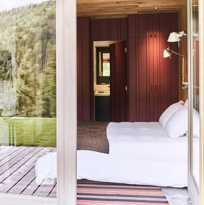 Vue depuis la terrasse en bois sur une chambre d'hôtel chalet avec lambris bordeaux, lit double en linge blanc, liseuse design dorée et tapis à rayures. La baie vitrée reflète la prairie et la forêt environnantes. Salle de bain visible en enfilade. Design alpin contemporain et coloré.