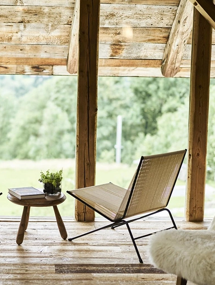 Fauteuil lounge design à structure métallique noire et assise tressée beige, posé sur un plancher en bois ancien dans un chalet alpin. Petite table basse en bois avec un livre et une plante verte. Grande baie vitrée donnant sur une prairie et des arbres en arrière-plan.